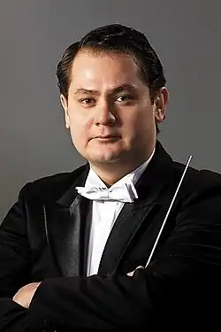 caucasian male with beard and square spectacles, wearing a tightly-buttoned tuxedo, acting as master of ceremonies