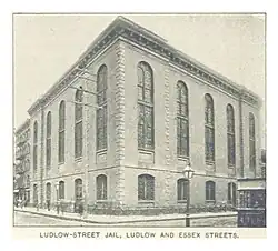 This is a black-and-white photo of the Ludlow Street Jail, a cuboid brick building visible here on two sides. Ten arched windows, five large and five small, are each on both sides. A sidewalk wraps around the building. The photo is originally captioned in all-caps, "Ludlow Street Jail, Ludlow and Exeter Streets".
