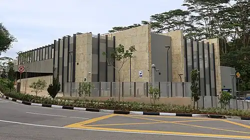 The station entrance located at the street level. The structure is a rectangular beige building with stripes of grey and black cladding.