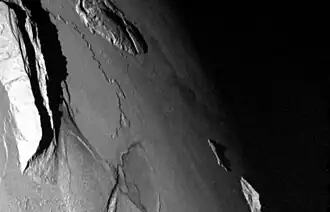 A portion of a planetary body with a pair of large, mountainous ridges on the left side of the image, a shorter, rugged domical mountain at top center, an elliptical pit near bottom center, and the boundary between the dayside (to the left) and the nightside (to the right) running down the right side of the image. Two small mountain peaks are seen near this boundary at lower right.