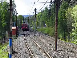 A double-track railway surrounded by trees runs down the middle of the image. On the left track is worn-looking red train. Above the tracks are overhead wires.