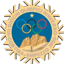 A stylized snowflake with the Olympic rings, a star and mountains. Surrounding the perimeter of the snowflake are the words, "VII Giochi Olimpici d'Inverno, Cortina d'Ampezzo 1956"