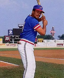 A man wearing a blue baseball jersey with red and white trim, a blue cap with a white "N" on the center, and white pants stands on a baseball field posed with bat, ready to swing.