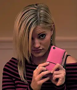 iJustine, tilting her head forward obscuring one of her eyes with her hair, is showing her iPhone, with a pink covering the back.
