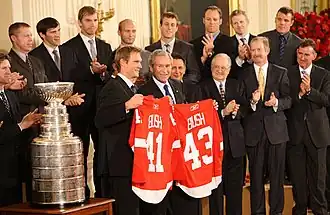 In the front row, Nicklas Lidstrom holds a Red Wings jersey with the last name "BUSH" and the number "41" while George W. Bush holds a Red Wings jersey with the last name "BUSH" and the number "43". To Lidstrom's right is the Stanley Cup. Second row, left to right: Mike Babcock, Ken Holland, Jim Devellano, Jim Nill, unknown man. Third row, left to right: Kris Draper, Pavel Datsyuk, Mikael Samuelsson, Brian Rafalski, Derek Meech, Kirk Maltby, Chris Osgood, Aaron Downey.