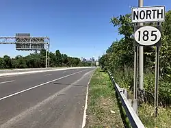 A road sign reading north above a black rectangle with a white oval containing 185 in black numerals. Trees and industrial buildings can be seen in the background.