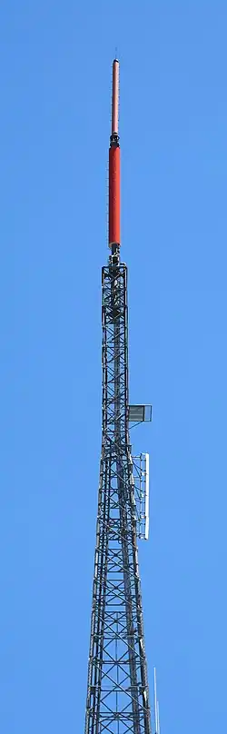 A gray lattice tower, set against a blue sky. A small pink cylindrical antenna and a larger red cylindrical antenna top the structure.