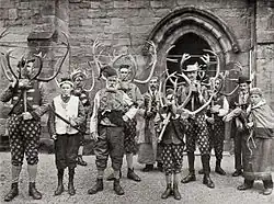 Black and white photograph of eleven men. Six carry reindeer horns. All but one are dressed in mock-medieval outfits; one wears a suit and bowler hat and carries a concertina