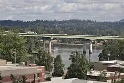 View of a highway bridge on four columns crossing a river with low-rise buildings in the background.