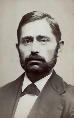 A young man in a suit with tightly-combed hair and a short, dark beard