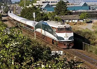 A passenger train with white, dark brown, and dark green stripes. The green stripe curves up along the side of the diesel locomotive.