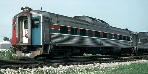 Stainless steel passenger rail cars with red and blue stripes horizontally across the windows. The visible end is covered with three vertical red, white, and blue stripes.