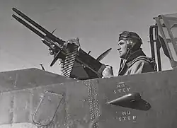 Black and white photograph of a man seated in an aircraft cockpit holding the rear handles of a large pair of guns