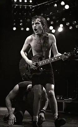 This photo is in black and white. Angus, aged 27, is shown playing guitar with both hands. He is seen shirtless, with shorts, and the socks-shoes combo. Brian Johnson, aged 34, is shown behind Angus, with his hands and knees on the ground. He is seen wearing an Irish hat, a tank top, jeans and shoes. Both are shown in right profile.