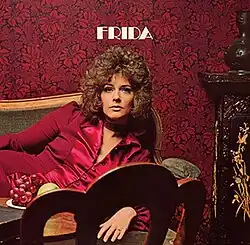 A curly-haired woman in a red suit and dress reclining on a chaise and facing forward. The text reads "FRIDA".