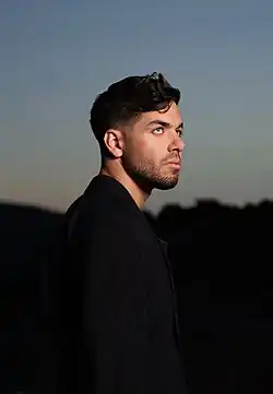 Profile image of Matteo Milleri wearing dark clothing, looking up and to the right in front of a blurry twilit background