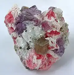 An apatite crystal sits front and center on cherry-red rhodochroite rhombs, purple fluorite cubes, quartz and a dusting of brass-yellow pyrite cubes.