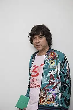 Rainer Ganahl wearing a white print t-shirt and colourful jacket, holding what appears to be an electronic table in right hand leaning against a wall and looking directly at camera