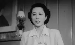 An attractive young Japanese woman, wearing a white blouse, is shown talking, photographed from below; a lamp, some bottles on a mantlepiece and part of a painting are visible in the left background.