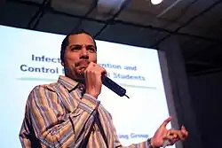 A person holds a microphone. They are standing in front of a projector displaying a PowerPoint slide.