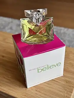 Glass octahedron perfume bottle containing green liquid sits atop the white, pink, and dotted retail cube box for Believe by Britney Spears