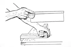 The workpiece is held against the fence of a bench hook.