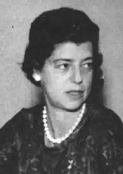 A white woman with dark coiffed hair, wearing a dark print dress and white beads with matching earrings