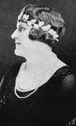 A woman with light skin and dark hair, photographed in profile, wearing a laurel-leaf headband low over her brow, and a dark scoop-necked gown, and two strands of pearls