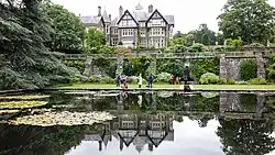 In the foreground is a lily pond, with a terrace wall behind, and in the background a substantial half-timbered country house surrounded by mature trees