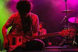 Two men perform on a stage, next to a drum kit and microphone. The man in the foreground rests on one knee, playing a guitar.