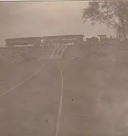 A small steam locomotive and tender, along with two passenger cars, parked in front of an open field. Stairs connect the field to the railroad tracks, which are at a higher elevation.