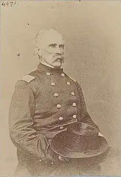 Sepia toned photo shows a seated man with a bushy moustache. He wears a dark military uniform and holds his hat in his lap.