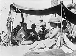 A number of men dressed in shorts and shirts with sleeves rolled up, one in a singlet sit in the shade of an awning. Several wear pith helmets, one of whom sits in the sun shirtless outside the awning leaning against a pole holding up the awning.