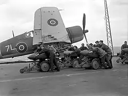 Black and white photograph of a group of men pushing bombs on trolleys on the deck of an aircraft carrier at sea. A single-engined aircraft is located immediately behind the men.
