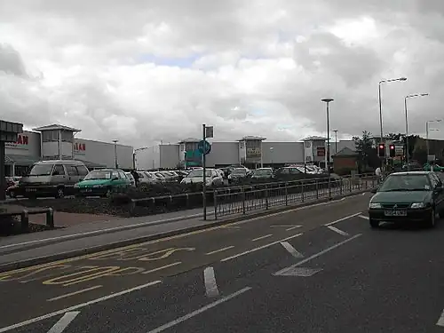 Modern buildings at rear with many parked cars beside a main road with bus stop and a traffic light controlled pedestrian crossing on a dull cloudy day