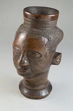 Head goblet (Mbwoongntey); 19th century; wood; Brooklyn Museum. It has a one-inch cylindrical lip with linear decoration. The hair is made up of crosshatched lines with a raised diamond-shaped segment on the back of the head. Its cheeks have curved multilinear scarification.