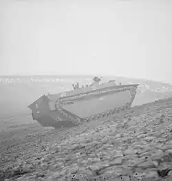 A tracked armoured vehicle drives up the bank of a river. In the background lies the wrecked frame of a metal bridge.