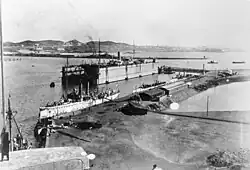 A black and white photograph of a large ship, along with two other small ships, docked inside the break waters of a harbor or port