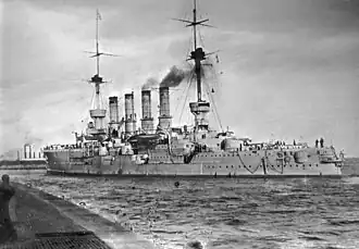 A large ship lies close to a wharf; dark gray smoke drifts up from its four tall smokestacks