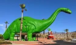 Two bright green concrete dinosaur sculptures in desert setting, with T-Rex wearing painted Santa suit