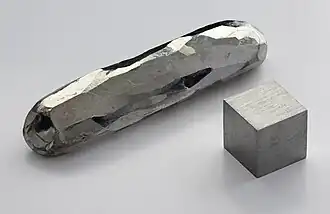 A bar and a cube of a silvery metal crystal lying on a grey surface
