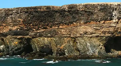 Cliff at Caleta Negra, Ajuy on Fuerteventura. The lower third of the cliff consists of steeply-tilted pre-Miocene oceanic sedimentary rocks. These rocks are unconformably overlain by Pliocene light-coloured cross-bedded beach sedimentary rocks, black pillow lava, and a black lava flow.[301][302] (Scale: cliff height is approximately 40&nbsp;m (130&nbsp;ft)[303])