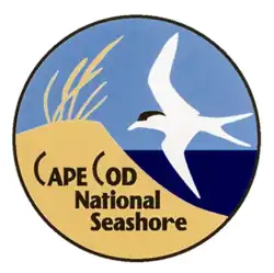 A circular logo depicting a sand dune topped with beach grass next to the ocean, with a white sea bird flying overhead.