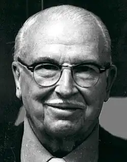 Black and white publicity photograph of a smiling, elderly, bespectacled man, with short, receding white hair and a narrow, white moustache.