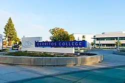 Sign for college with Cerritos College logo of a C within a C. Sign is in a pedestrian plaza with buildings in background.