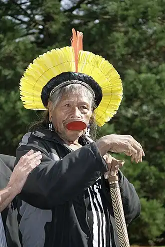 An elderly man is looking forward. He wears traditional garments on his head and lower lip