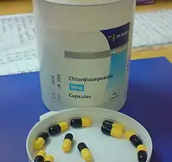 White bottle on blue pad atop a desk. The bottle cap is off and is upside down on the pad in front of the bottle. In the cap are a dozen black-and-yellow capsules.