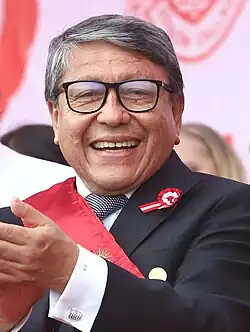 Image of man in the foreground with glasses and grey hair, smiling and clapping. He is wearing a black suit coat with a red sash.