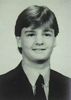 A monochrome portrait photo of a white teenaged boy, he is wearing a dark suit and tie, smiling while facing and looking slightly to the camera's right