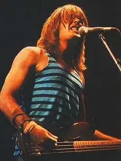 Williams, about 31, shown in upper body shot. He sings into microphone on a stand while strumming his guitar. He wears a singlet top and has shoulder length hair.